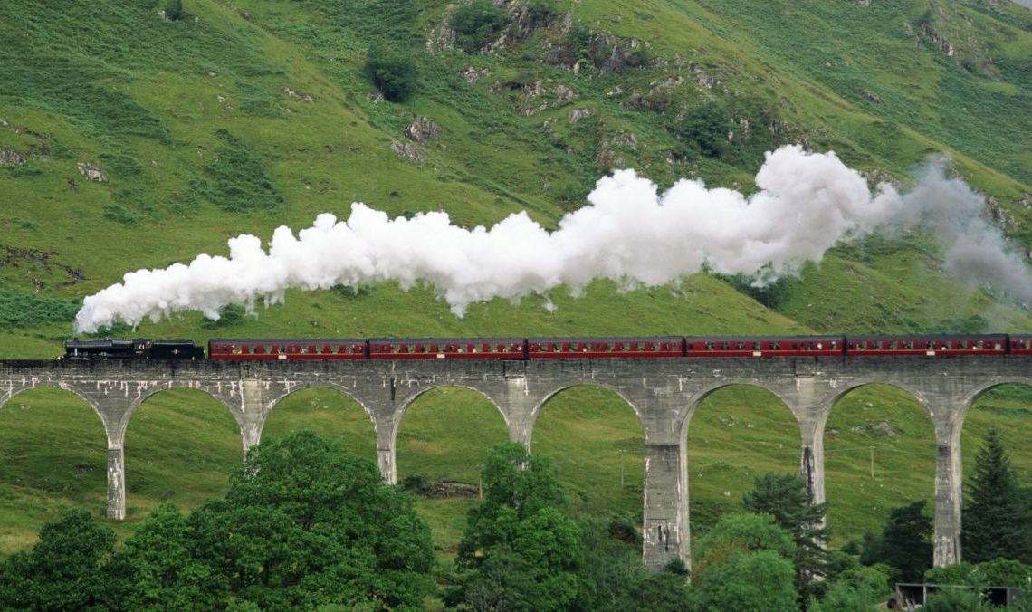 The Flying Scotsman train goes over a bridge, lots of steam coming from its funnel.